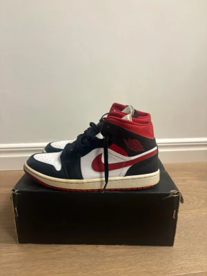 Nike Air Jordan 1 Retro High OG - Säljer ett par Nike Air Jordan 1 Retro High OG sneakers i svart, vitt och rött. Skorna har klassisk hög siluett, svart läder på tå och sidor, vit tåbox och paneler samt röda detaljer på swoosh och krage. Ikonisk Air Jordan-logga på sidan och svart snörning.