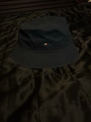 Mörkblå bucket hat Tommy Hilfiger - Snygg mörkblå bucket hat från Tommy Hilfiger med den klassiska lilla loggan framtill. Hatten är i mjukt tyg och har en avslappnad passform med bred brätte. Perfekt accessoar för en trendig streetwear-look.