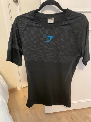 Gymshark onyx 5.0 - Svart kortärmad träningströja från Gymshark med blå logga på bröstet. Tillverkad i ett lätt och ventilerande syntetmaterial med mönstrad ovandel och slät nederdel. Perfekt för intensiva träningspass och gym.