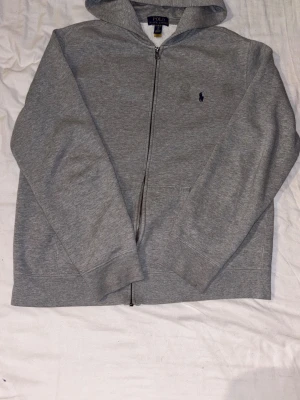 Grå hoodie från Polo Ralph Lauren - Snygg grå hoodie från Polo Ralph Lauren med dragkedja framtill och klassisk blå logga på bröstet. Tillverkad i mjukt bomullsmaterial och har en huva samt fickor på sidorna. Perfekt för en chill och stilren look.