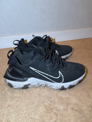 Nike React Element 55 svarta sneakers - Svarta Nike React Element 55 sneakers med vit sula och vit swoosh på sidan. Ovandelen är i mesh och syntetmaterial, med svarta skosnören och detaljer. Sulan har en unik design med svarta och vita partier. Perfekta för en sportig och trendig look.