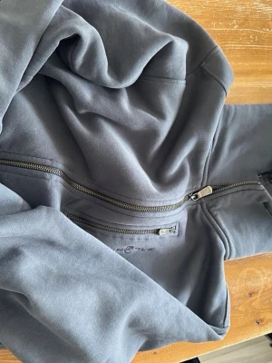 Grå hoodie från Sail Racing - Snygg grå hoodie från Sail Racing med dragkedja framtill och två praktiska bröstfickor med dragkedjor. Broderad logga på bröstet och mjukt material som känns skönt mot huden. Perfekt för en avslappnad och sportig stil.
