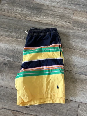 Randiga badbyxor från Polo Ralph Lauren - Säljer ett par färgglada badbyxor från Polo Ralph Lauren med breda ränder i marinblått, gult, grönt och orange. Klassisk passform med dragsko i midjan och broderad logga på benet. Perfekta för stranden eller poolen! Bredd är 39 och längd 44