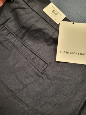 Mörkgrå chinos från These Glory Days 54 L - Snygga mörkgrå chinos från These Glory Days i storlek 54. Klassisk rak passform med diskreta fickor och bälteshällor. Tillverkade i ett mjukt och slitstarkt bomullsmaterial som ger en clean och stilren look. Perfekta för dig som gillar enkel och modern stil.