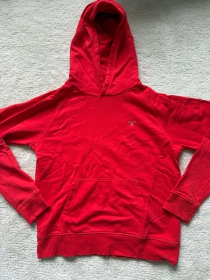 Röd hoodie från GANT med huva - Snygg röd hoodie från GANT med klassisk huva och känguruficka framtill. Liten GANT-logga broderad på bröstet. Tillverkad i mjuk bomullsmix som känns skön mot huden. Perfekt för en chill och avslappnad stil.