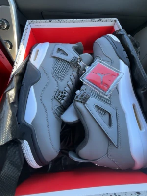 Air Jordan 4 Retro Cool Grey - Snygga Air Jordan 4 Retro Cool Grey sneakers med grå ovandel i läder och mesh, vita och svarta detaljer samt gulddetalj vid sulan. Klassisk Jumpman-tag i plast hänger vid snörningen. Perfekta för dig som vill sticka ut med stilrena och ikoniska sneakers.