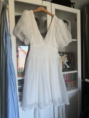 Vit kort omlottklänning med puffärm - Supersöt vit kort klänning med djup v-ringning, stora puffärmar och omlottdetalj i midjan som knyts bak. Tillverkad i ett lätt, skirt tyg med struktur som ger en luftig känsla. Perfekt för sommarens alla tillfällen!
