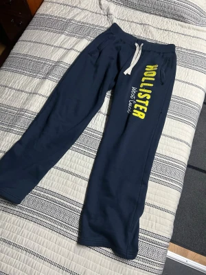 Mörkblå mjukisbyxor Hollister - Mörkblå mjukisbyxor från Hollister med stort gult tryck längs benet där det står 'HOLLISTER West Coast'. Byxorna har vita dragsnören i midjan och två sidofickor. Tillverkade i mjukt och bekvämt material, perfekta för chill dagar.