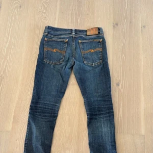 Blå jeans från Nudie Jeans - Säljer ett par klassiska blå jeans från Nudie Jeans med smal passform. Jeansen har snygga slitningar, kontrastsömmar och ikoniska broderade bakfickor. Tillverkade i denim med läderpatch i midjan. Perfekta för en avslappnad och trendig look.