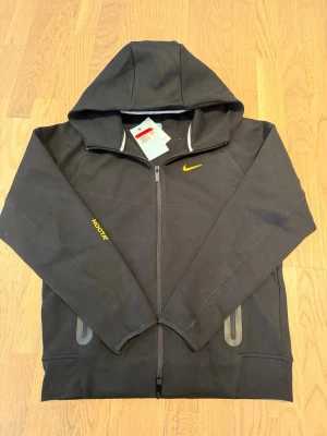 Nike NOCTA hoodie - Svart hoodie från Nike NOCTA-kollektionen med gul logga på bröstet och NOCTA-tryck på ärmen. Hoodien har hel dragkedja, två fickor framtill och huva. Materialet är mjukt och bekvämt, perfekt för en avslappnad streetwear-look.