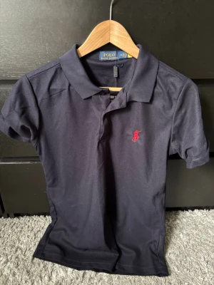 Mörkblå pikétröja från Polo Ralph Lauren som passar S - .Klassisk blå pikétröja från Polo Ralph Lauren med korta ärmar och krage. Tröjan är storlek M men passar storlek S! Tröjan är nytt skick och inga skador, pris kan diskuteras. Hör av er vid funderingar!