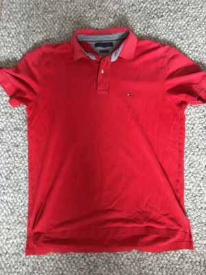 Röd pikétröja från Tommy Hilfiger - Klassisk röd pikétröja från Tommy Hilfiger med liten logga på bröstet. Tröjan har krage, knappar vid halsen och korta ärmar. Perfekt för en avslappnad men ändå stilren look.