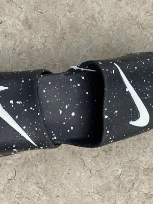 Svarta Nike tofflor med vita stänk - Säljer ett par svarta Nike tofflor med vita färgstänk och den klassiska vita Swoosh-loggan på ovansidan. Tofflorna har en bred rem och är tillverkade i syntetmaterial, perfekta för chill dagar eller efter träningen.
