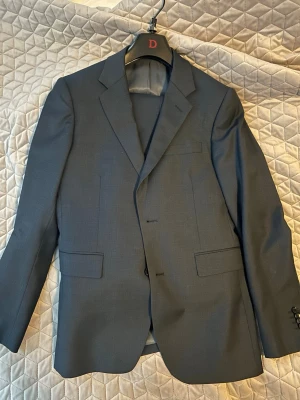 Mörkblå kostym från dressman - Snygg mörkblå kostym från Dressman med klassisk kavaj och matchande byxor. Kavajen har två knappar, fickor med lock och innerfickor. Kostymen är helfodrad och har en stilren, tidlös look som passar perfekt till mer uppklädda tillfällen såsom balen. Jag säljer denna kostym då jag använt den en gång vilket var på min bal i 9an och nu är den för liten 10/10 skick på kostymen