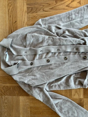 Beige kofta från Brown Avenue - Snygg beige kofta från Brown Avenue med knappar framtill och en bröstficka. Koftan har klassisk krage och är tillverkad i mjukt stickat material. Perfekt att slänga över en t-shirt eller topp för en chill look.