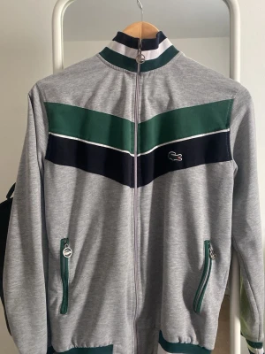 Grå zip tröja från Lacoste - Snygg grå ziptröja från Lacoste med gröna och marinblå detaljer samt vita ränder. Jackan har hel dragkedja, två fickor med dragkedja och klassisk Lacoste-logga på bröstet. Perfekt för dig som gillar sportig och retro stil.