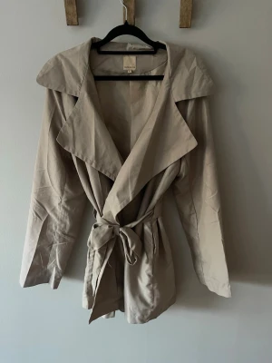 Beige trenchcoat med knytskärp - Supersnygg kort beige trenchcoat från Jeane Blush med knytskärp i midjan. Lite längre passform och tunnare tyg som gör den perfekt för lite varmare väder. 