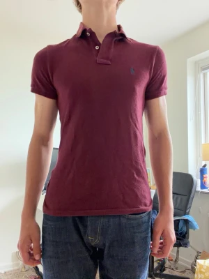 Vinröd pikétröja från Ralph Lauren - Snygg vinröd pikétröja från Ralph Lauren med klassisk krage, två knappar och den ikoniska broderade loggan på bröstet. Tröjan har korta ärmar och är gjord i mjukt bomullsmaterial. Perfekt till jeans eller chinos för en clean look.