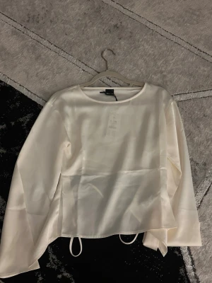Vit satinblus med knytband - Stilren vit blus från Gina i satintyg. Blusen har rund halsringning, långa ärmar och dekorativa knytband i midjan för en trendig look. Perfekt att styla med jeans eller kjol för en chic outfit.