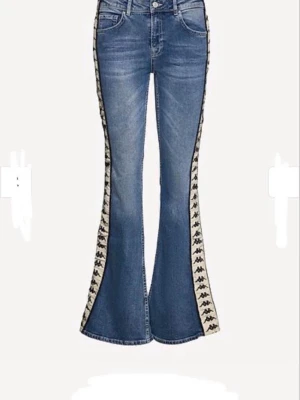 Blå utsvängda jeans från Kappa - Säljer ett par blå utsvängda jeans från Kappa med ikoniska vita sidoränder och svart logga längs benen. Jeansen har klassisk femficksmodell och hög midja. Materialet är denim med stretch för extra komfort. Perfekta för dig som vill sticka ut med retrovibbar!