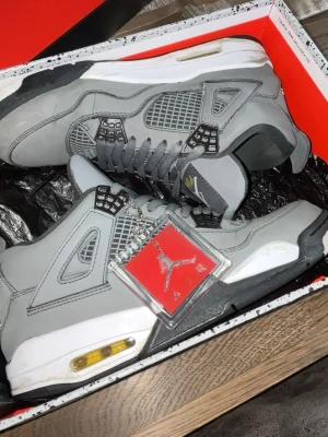 Air Jordan 4 Cool Grey - Grymma Air Jordan 4 Cool Grey med grå mocka, svarta och vita detaljer samt gula accenter vid sulan. Klassisk Jumpman-logga på tungan och hängande tag. Perfekta sneakers för dig som gillar streetwear och ikonisk design.