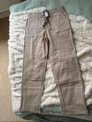 Beige mjukisbyxor med raka ben (Stella Low Waist)  - Säljer ett par beige mjukisbyxor med raka ben och bred resår i midjan. Byxorna har stora fickor framtill och en bakficka, samt dragsko i midjan för skön passform. Perfekta för chill dagar eller när du vill vara extra bekväm.