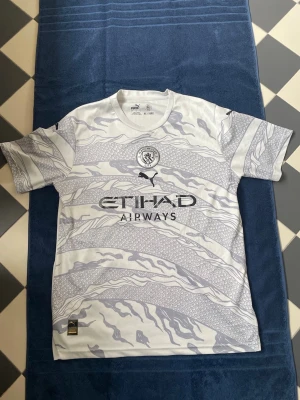 Manchester City bortatröja Puma - Snygg Manchester City bortatröja från Puma i vitt med ljusgrått mönster och klubbmärke på bröstet. Tröjan har korta ärmar, rund hals och Etihad Airways-logga framtill. Tillverkad i lätt och ventilerande polyester, perfekt för fotboll eller träning. Texten är lite urtvättad. 