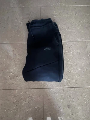 Svarta Nike mjukisbyxor med dragsko - Säljer ett par svarta mjukisbyxor från Nike med smal passform och ribbade muddar vid bensluten. Byxorna har en justerbar dragsko i midjan och diskret Nike-logga på benet. Perfekta för chill dagar eller träning.