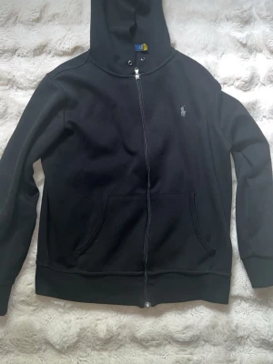 Svart zip från Polo Ralph Lauren - Klassisk svart hoodie från Polo Ralph Lauren med dragkedja framtill och broderad logga på bröstet. Hoodien har huva med dragsko och känguruficka. Tillverkad i mjukt bomullsmaterial som är skönt att bära. Har inga skador eller defekter 