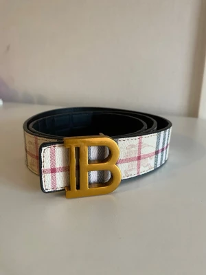 Rutig  bälte med B-spänne - Snyggt bälte  med klassiskt rutigt mönster i vitt, rött och grått. Bältet har ett stort guldfärgat B-spänne framtill och är gjort i skinn. Perfekt accessoar för att lyfta din outfit.