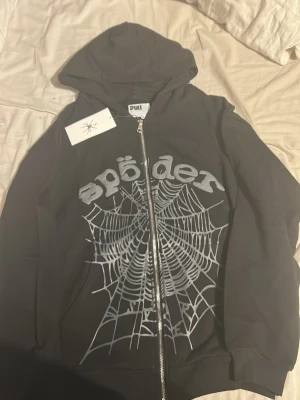 Sp5der hoodie - Säljer en svart hoodie från Sp5der med dragkedja och stor huva. På framsidan finns ett stort tryck med texten 'sp5der' och ett detaljerat spindelnät i vitt. Hoodien har ribbade muddar och är perfekt för dig som gillar streetwear och grafiska prints.