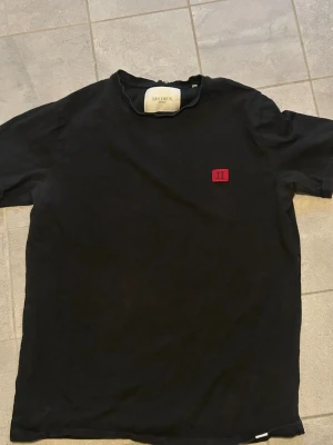 Svart t-shirt från Les Deux - Snygg svart t-shirt från Les Deux med korta ärmar och en liten röd patch med logga på bröstet. Tillverkad i mjuk bomull för skön känsla. Enkel och stilren design som passar till det mesta.