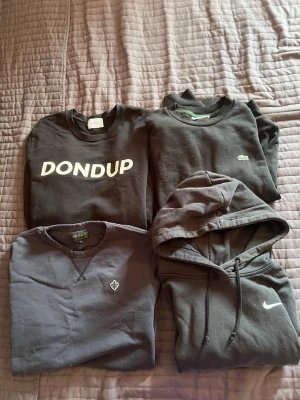 Svarta sweatshirts från Nike, Lacoste, Dondup - Fyra svarta sweatshirts i olika modeller: en hoodie från Nike med vit logga, en crewneck från Lacoste med liten grön krokodil, en Dondup-tröja med stor vit logga och en Morris-tröja med broderad logga. Alla har klassisk passform och rund hals.