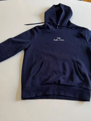 Mörkblå hoodie från Polo Ralph Lauren - Snygg mörkblå hoodie från Polo Ralph Lauren med broderad logga på bröstet och ärmen. Klassisk känguruficka framtill och justerbar huva med dragsko. Tillverkad i mjukt bomullsmaterial som är skönt att bära. Perfekt för en avslappnad och stilren look. Endast andvänd ett fåtal gånger med ett mycket bra skick! 