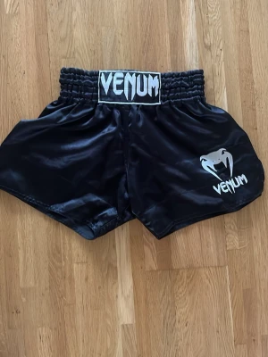 Muay Thai-shorts Venum - Snygga svarta Muay Thai-shorts från Venum med vitt logotryck och resår i midjan. Tillverkade i glansigt, lätt och slitstarkt material som ger bra rörelsefrihet. Perfekta för kampsportsträning och sparring inom boxning, mma, muay thai mm. 