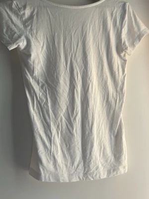 Vit t-shirt med djup urringning - En basic vit t-shirt i mjuk bomull med korta ärmar och en djup, rundad urringning i ryggen. Perfekt att styla till jeans eller kjol för en clean och enkel look. Passformen är normal och tyget är tunt och följsamt.