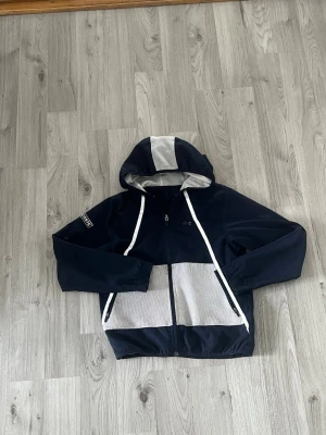 Marinblå vindjacka från Lacoste - Snygg marinblå vindjacka från Lacoste med vita meshdetaljer på huva och fickor. Jackan har dragkedja framtill, två fickor med dragkedja och justerbar huva med vita snören. Lacoste-logga på ärmen ger en sportig vibe. Perfekt för blåsiga dagar.