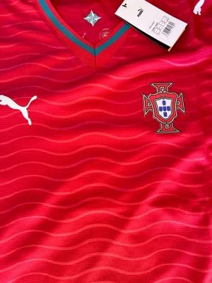 Portugal fotbollströja från Puma - Röd Portugal fotbollströja från Puma med vågigt mönster, broderad landslagssköld på bröstet och vit Puma-logga. V-ringad krage med gröna och röda detaljer. Tillverkad i lätt och ventilerande material, perfekt för träning eller match.