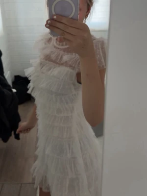 Vit volangklänning med meshdetaljer - Supersöt vit kortklänning med flera lager volanger och transparenta meshpartier upptill. Klänningen har korta ärmar och en lätt v-ringad rygg. Perfekt för dig som vill ha en romantisk och lekfull look.
