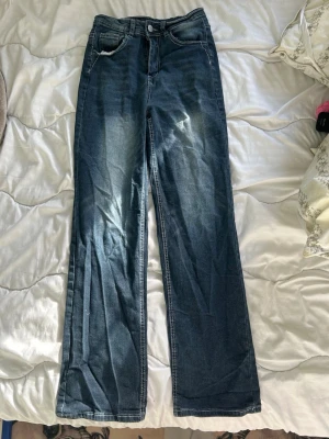 Blå raka jeans med hög midja - Säljer ett par klassiska blå jeans med raka ben och hög midja. Jeansen har fem fickor, knappgylf och subtila slitningar framtill för en avslappnad look. Perfekta till en trendig vardagsstil.