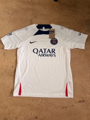 Vit PSG träningströja från Nike - Säljer en vit PSG matchtröja från Nike med mörkblå detaljer och röda inslag vid sidorna. Tröjan har PSG-logga på bröstet, Qatar Airways-tryck och diskret mönster på ärmarna. Perfekt för fotbollsträning eller att bära till vardags.