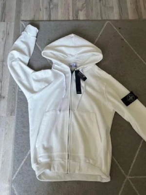 Vit hoodie från Stone Island - Snygg vit hoodie från Stone Island med dragkedja framtill och klassisk huva. Hoodien har en stor ficka på magen och den ikoniska Stone Island-loggan på vänster ärm. Tillverkad i mjukt bomullsmaterial för skön komfort.