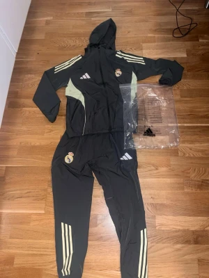 Real Madrid windbreaker träningsdress Adidas - Snygg svart träningsdress från Adidas x Real Madrid med ljusgula detaljer och klubbmärke på både jacka och byxor. Setet har huva, dragkedja och klassiska Adidas-ränder på ärmar och ben. Perfekt för fotbollsträning eller chill.