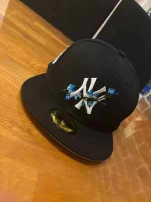 Svart New Era NY keps med blommor - Snygg svart keps från New Era med broderad vit NY-logga och blå blommor på framsidan. Klassisk flat peak och New Era-logga på sidan. Perfekt för dig som vill ha en unik och trendig accessoar till din streetwear-look.