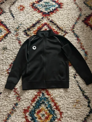 Svart träningsjacka från Craftpassar både för storlek S, M och L - Svart träningsjacka från Craft med dragkedja framtill och ståkrage. Jackan har två sidofickor och en vit logotyp på bröstet. Tillverkad i ett slätt, sportigt material som passar perfekt till träning och uppvärmning. passar både för storlek S, M och L
