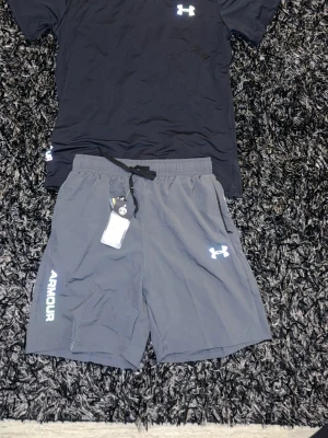 Grå träningsshorts från Under Armour - Snygga grå träningsshorts från Under Armour med vit logga på benet och texten 'ARMOUR' längs sidan. Shortsen har elastisk midja med snörning och är tillverkade i ett lätt och snabbtorkande material som passar perfekt för träning.