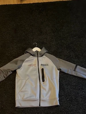Grå hoodie från Hoodrich - Snygg grå hoodie från Hoodrich med dragkedja framtill och svart tryck på bröstet. Ärmarna är i en mörkare grå nyans och huvan har tryck. På ryggen står det 'Hoodrich Lifestyle' i svart och rött. Perfekt för en streetwear-look.