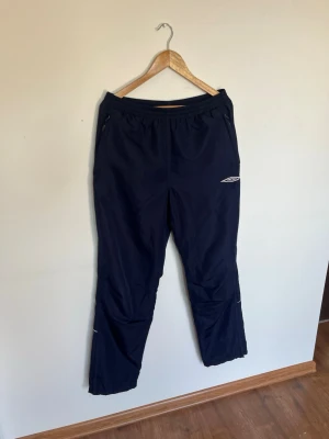 Retro Mörkblå joggers från Umbro - Säljer ett par mörkblå joggers från Umbro med resår i midjan och dragkedjor vid fickorna. Byxorna har meshfoder på insidan för extra komfort och är tillverkade i ett lätt, snabbtorkande material. Perfekta för träning eller uppvärmning. Vintage look. Storlek M. Fint skick