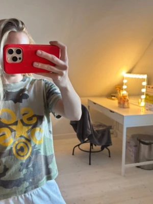 Camouflage t-shirt med gul print - Säljer en croppad t-shirt med camouflage-mönster i gröna och bruna toner. Framtill finns ett stort gult tryck med stjärna och bokstäver. T-shirten har korta ärmar och rund hals. Perfekt för en avslappnad och cool stil.         passar till s\m 