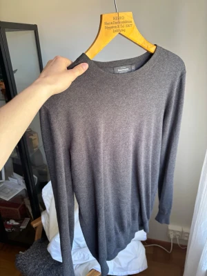 Mörkgrå tunn tröja från Dressmann - Snygg och enkel mörkgrå tröja från Dressmann i tunt, mjukt material. Tröjan har rund halsringning och långa ärmar, perfekt för lager på lager eller som den är. Passar till många olika stilar och är riktigt bekväm.